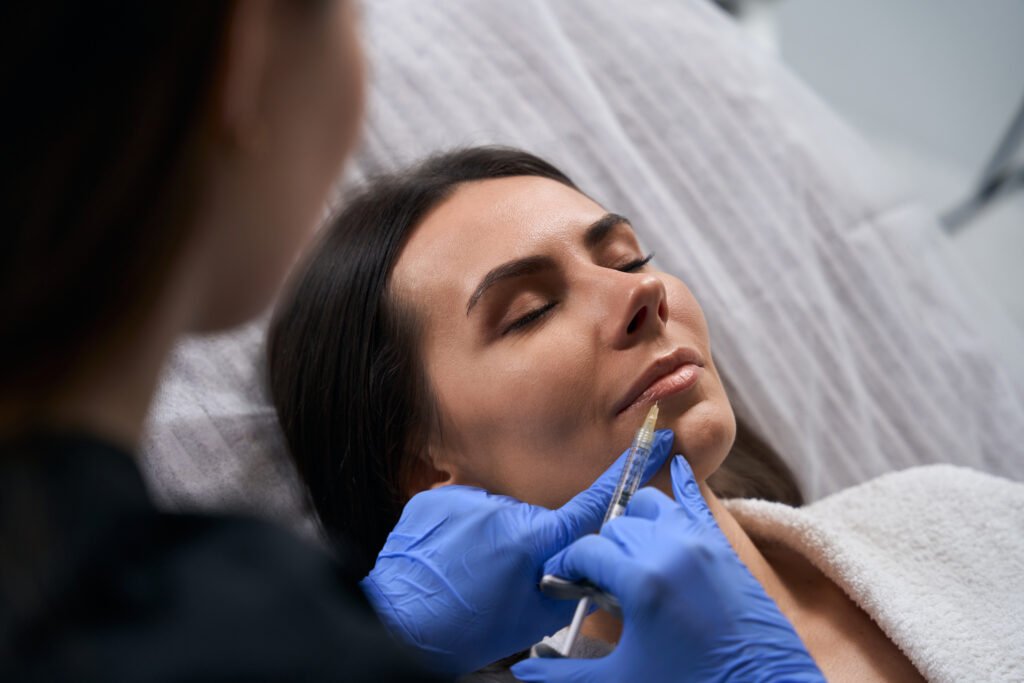 Cosmetologist in medic coat and gloves holding syringe and making beauty injections for face to client